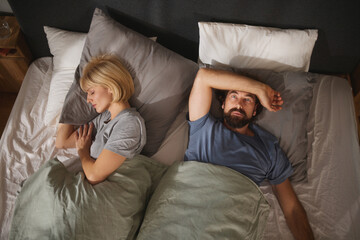 Two people lie in bed, turned away from each other. The room is dimly lit, creating a tense atmosphere as they struggle with their relationship issues and emotional distance.