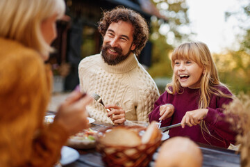 A family enjoys a joyful weekend in autumn, sitting outdoors at a wooden table filled with food, sharing laughter and a warm connection while dressed in cozy sweaters.