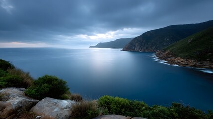 A dramatic moody twilight view of a serene sea inlet with a curved rocky coastline under cloudy skies