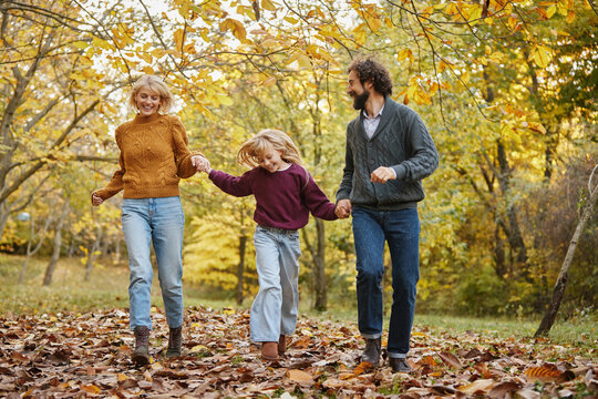 A family of three walks hand in hand through a vibrant park filled with colorful autumn leaves. They smile and share joyful moments on a sunny weekend. - Powered by Adobe