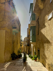 Atmospheric view of a typical narrow, stepped street in the historic city of Valletta, Malta. Strong backlighting highlights the traditional green Maltese balcony (gallarija) and the rustic limestone 
