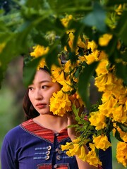 Portrait of Asian woman wearing traditional Thai dress standing near blooming yellow flowers in natural sunlight, symbol of beauty and local culture.