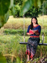 Asian woman in traditional Thai clothing sitting on swing reading book in rice field, relaxing rural scene representing peaceful countryside lifestyle.