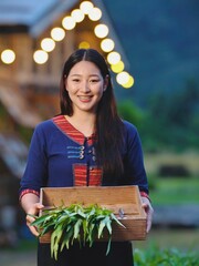 Smiling Asian woman in traditional Thai outfit holding wooden box with fresh vegetables, showing happiness and simplicity of rural lifestyle in countryside.