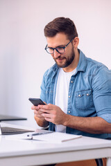 Young attractive man using mobile phone while working on laptop in his office.