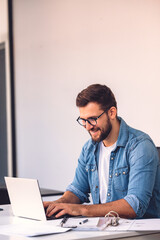 A smiling man with glasses is using a laptop in the office while sitting at his desk.