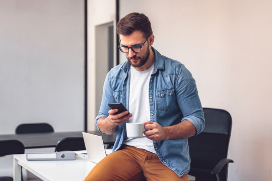 Handsome bearded man leaning on office desk, drinking coffee and looking at phone. - Powered by Adobe