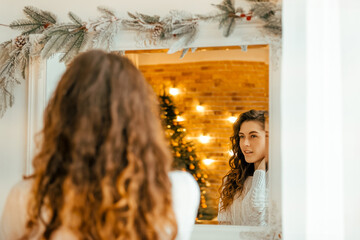 beautiful curly girl in a white sweater with dark hair sits in front of a mirror. portrait on the background of New Year's decor and a white window