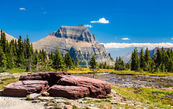 Scenic view of Mount Oberlin from Logan Pass in Glacier National Park. Red argillite rocks are in the foreground, next to an alpine stream and pine trees, under a clear blue sky