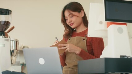 Smiling Young Asian woman small business owner working in cafe, using laptop for planning budget, cost and managing stock inventory at coffee shop. Female shop owner bookkeeping, checking bill at shop