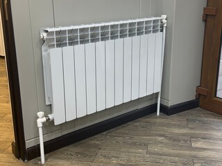 Modern white aluminum heating radiator installed in the corner of a gray wall and wood-like floor. The image highlights home comfort, heating systems, and interior design. Heat, winter, house.