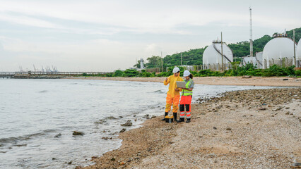 Two engineers, one in a yellow protective suit, inspect a coastal industrial site. They use a laptop and testing equipment on the beach near large gas storage tanks.