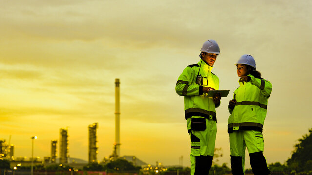 male and female engineer in high-vis gear and hard hats use a tablet. They are collaborating at an industrial factory or refinery site during a golden sunset.