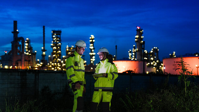 Petrochemical engineers in PPE conduct a night shift inspection at a refinery. They use a tablet for real-time data monitoring and auditing of the processing plant.