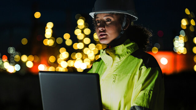 A close up of a female industrial worker in a hard hat and safety gear looking at a lit up factory or refinery at night. The background lights are blurred into bokeh.
