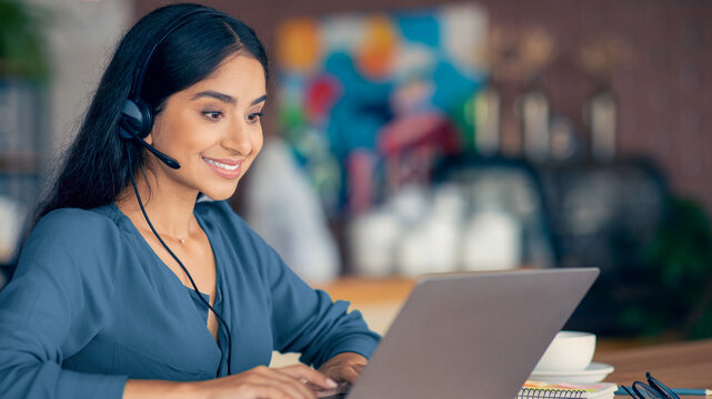 A young woman is focused on her laptop while wearing a headset in a lively cafe. She appears engaged and is likely participating in a virtual meeting or call. The background is colorful and dynamic.