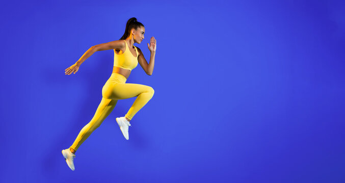 A female athlete in a vibrant yellow outfit is running energetically in mid-sprint. The background is a solid blue, highlighting her dynamic movement and athleticism.