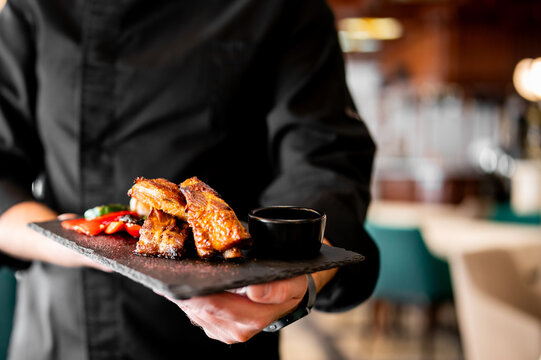 A chef holds a black slate platter featuring grilled pork ribs in BBQ sauce, garnished with red peppers and a dipping bowl, in a restaurant setting