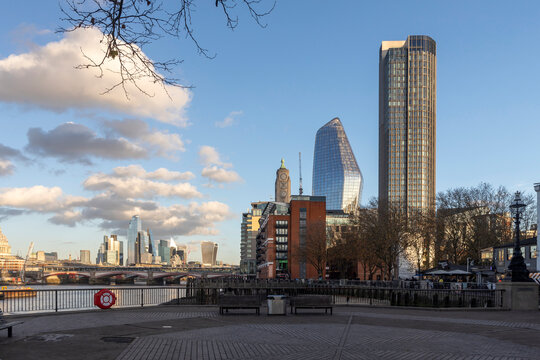 London, UK - December 1, 2024, A view of the city skyline from a paved riverbank, highlighting the juxtaposition of contemporary high-rise buildings and older structures under a bright, cloudy sky.