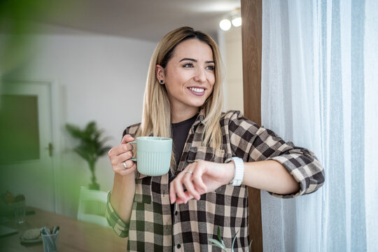 Young woman drinking coffee checking smartwatch at home