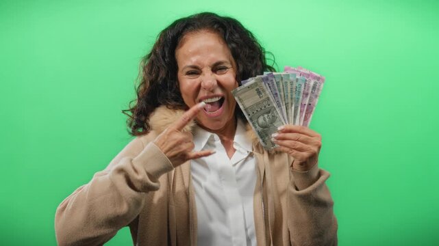 Middle age woman posing with indian rupee banknotes against a vibrant green backdrop, making a rock gesture showcasing confidence and financial empowerment. - Powered by Adobe