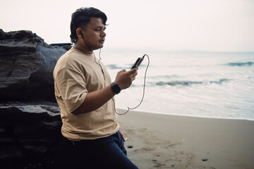 Cheerful Caucasian young man with smartphone weTropical mood. Stylish man relaxing on tropical beach on his holidays. Listening music, enjoying vacations, looking on horizon.