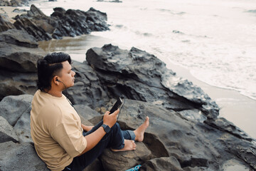 Cheerful Caucasian young man with smartphone weTropical mood. Stylish man relaxing on tropical beach on his holidays.  Listening music, enjoying vacations, looking   on horizon. 
