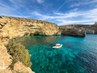 Idyllic high-angle view of a secluded bay on Comino Island, Malta, bathed in warm afternoon sunlight. The incredibly clear turquoise water is home to a small anchored motorboat, symbolizing summer rel