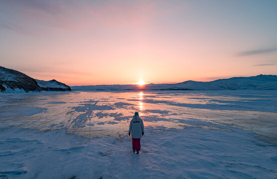Aerial view on the young woman walking on the blue cracked ice of Baikal at beautiful orange sunrise. Sun reflections on the ice. Winter landscape of frozen Baikal