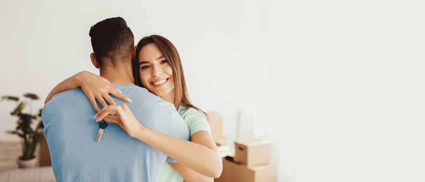 A happy couple shares a warm hug in their new home. The woman holds a house key, symbolizing their recent achievement, surrounded by moving boxes and a plant. - Powered by Adobe