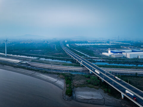 Aerial view of a long highway bridge with traffic crossing a river towards an industrial zone with wind turbines at dusk.