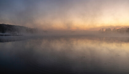 Misty lake at sunrise with gentle ripples reflecting the soft orange and purple sky and distant tree line
