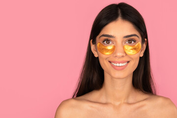 Smiling young woman wearing gold under-eye patches for skincare and rejuvenation. Beauty treatment concept for hydration and anti-aging on pink background