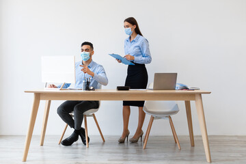 Young male boss and female colleague in face masks collaborate at a desk in a modern office. The manager points at the computer while the lady takes notes on a clipboard.
