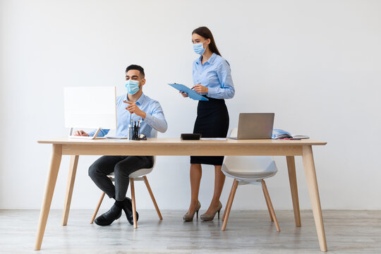 Young male boss and female colleague in face masks collaborate at a desk in a modern office. The manager points at the computer while the lady takes notes on a clipboard.