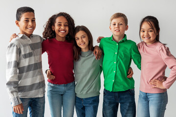 Obraz premium A group of cheerful multicultural preteen kids stands together, hugging and smiling at the camera. They wear colorful outfits and display a strong sense of friendship in a gray studio setting.