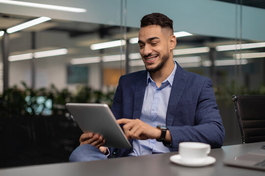 A millennial arabic businessman in a suit sits at a table in a contemporary office. He smiles as he checks a business app on his digital tablet. Morning light fills the space.