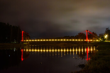 Illuminated suspension bridge glowing red at night, reflected beautifully in calm river water under a cloudy evening sky.