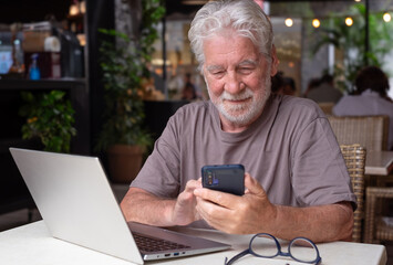Smiling bearded senior man sits in a cafe using laptop and smartphone. An elderly business man uses technology expressing confidence and contentment in a relaxed atmosphere.