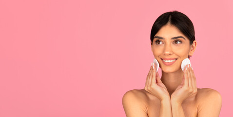 Smiling woman cleaning face with cotton pads on pink background. Studio portrait symbolizing daily...