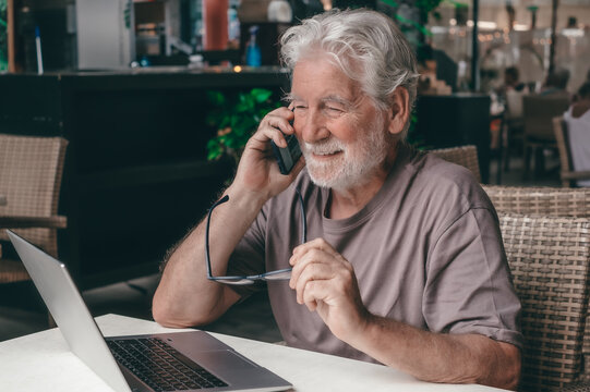 Smiling bearded senior man sits in a cafe using his laptop talking on smartphone. An elderly business man uses technology expressing confidence and contentment in a relaxed atmosphere.