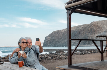Smilling senior couple sitting for a break with a cup of tea along the seacoast while using smartphone for a selfie. Serene retirement lifestyle