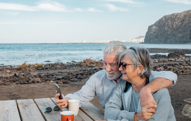 Smilling senior couple sitting for a break with a cup of tea along the seacoast while using smartphone in video call. Serene retirement lifestyle