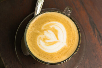 Top-down view of a latte with heart-shaped foam art in a glass cup on a dark wooden table.