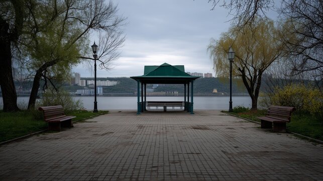 A serene riverside scene features a gazebo overlooking the calm water with benches placed nearby. Two benches sit along the paved path, surrounded by trees and soft grass.