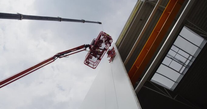 Construction crane hoists a large insulated sandwich panel, while workers on a boom lift secure it in place as a wall of a modern industrial building.