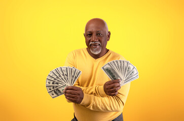 A joyful senior man smiles while holding two fans of cash, showcasing his excitement over a recent financial win. The bright orange background enhances the mood of celebration and prosperity.