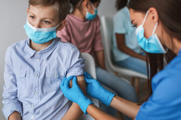 A preteen boy sits in a chair while a nurse administers the Covid-19 vaccine in a modern clinic. Other children wait in the background. The nurse gently applies a bandage on the boy's arm.