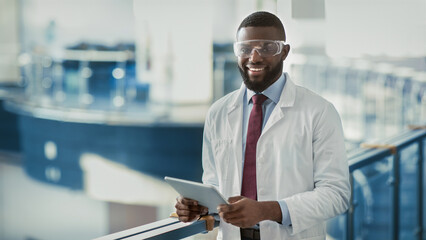 Happy young black man in workwear and protective glasses medical scientist with using digital tablet, analyzing data, posing at workplace, panorama with copy space. Jobs and occupations concept