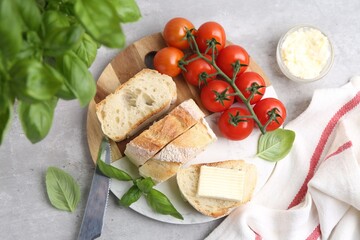 Slices of baguette with butter, basil, tomatoes and knife on light grey table, flat lay